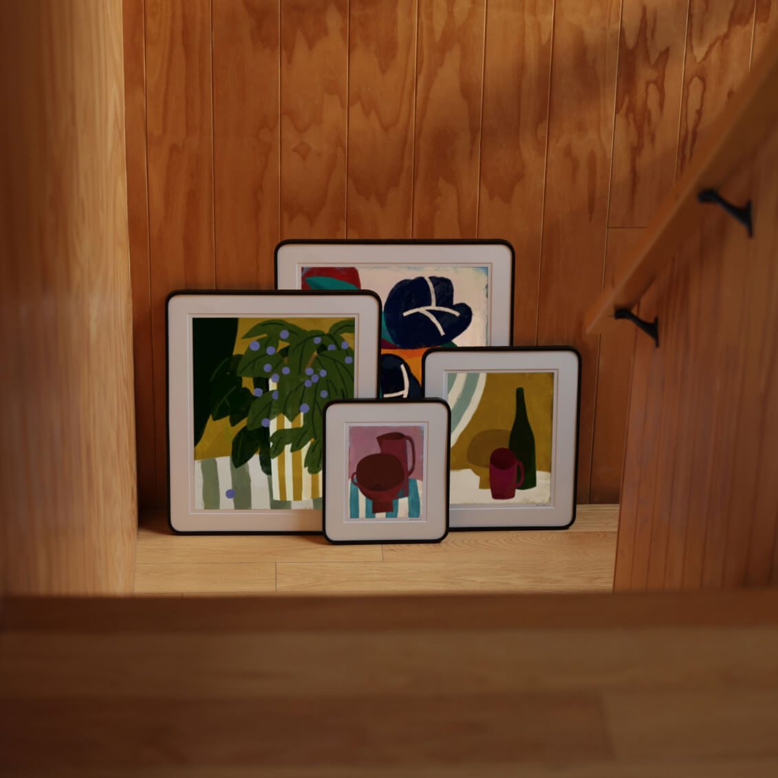 Set of framed artworks on a wooden shelf against a wooden wall.