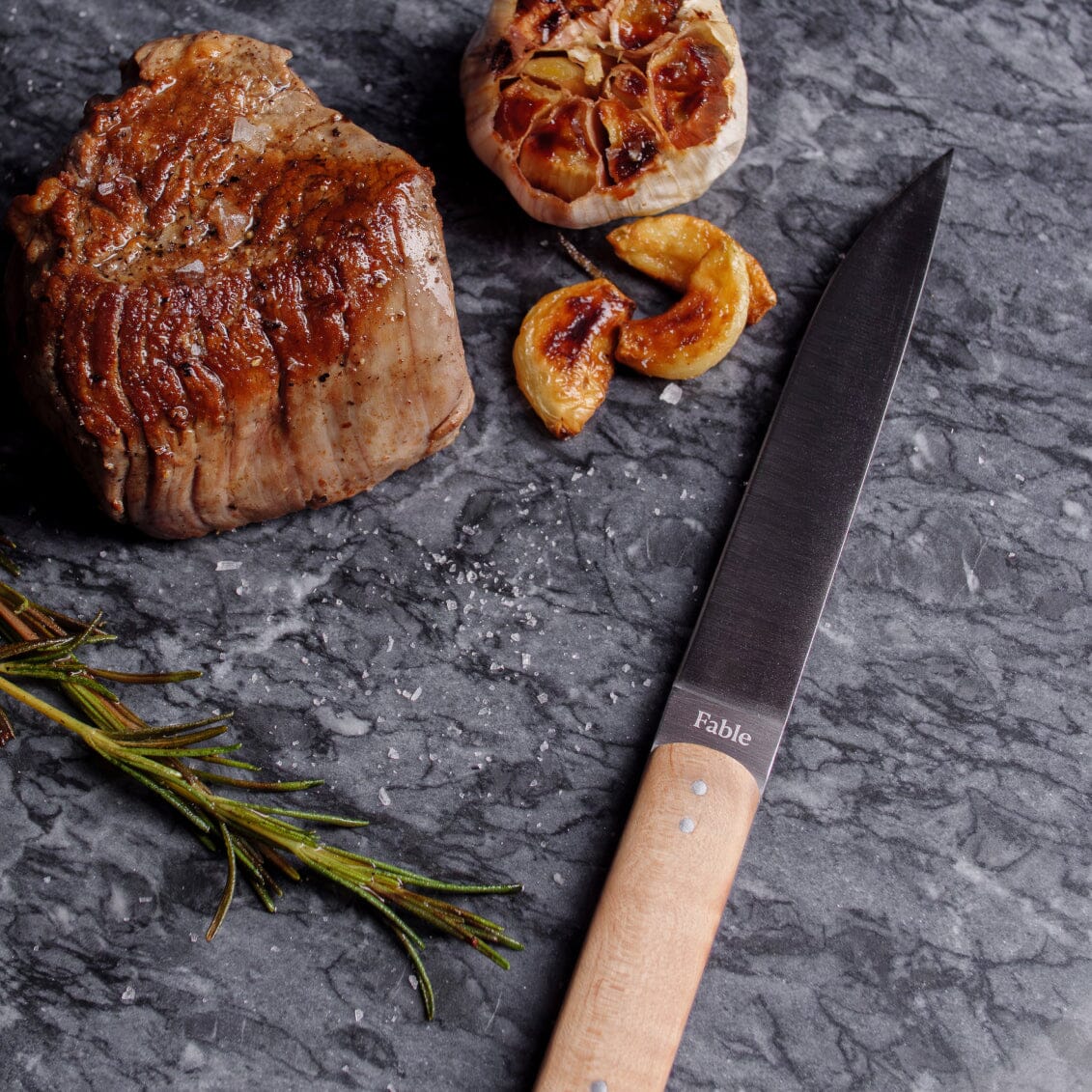 Steak, garlic, and a knife on a dark surface with 'Fable' branding.